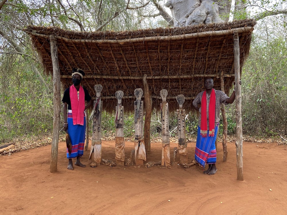 Two people wearing red and blue clothing stand on either side of five wooden sculptures under a shelter with a thatched roof. Two people wearing red and blue clothing stand on either side of five wooden sculptures under a shelter with a thatched roof.